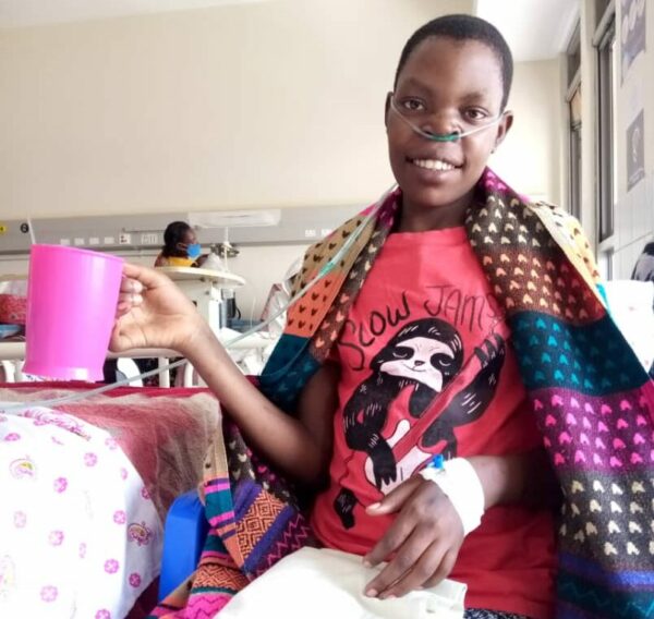 Girl with a blanket over her shoulders holding a pink water pitcher in a Ugandan hospital