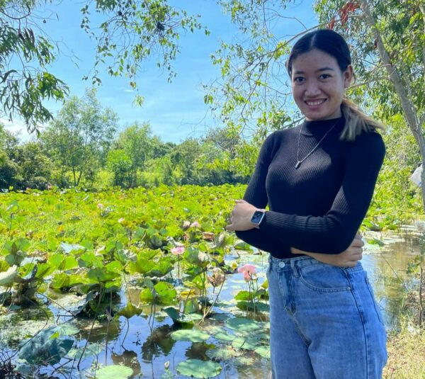 Young woman in a black top and jeans stands by a pond with water lilies in bloom