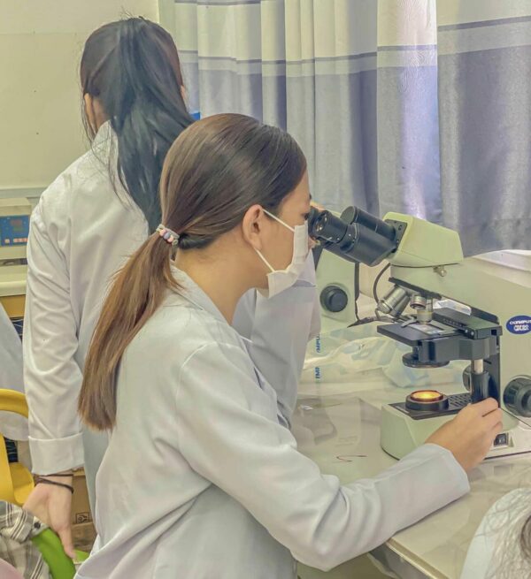 A young woman in a white lab coat and mask peers into a microscope in a lab.