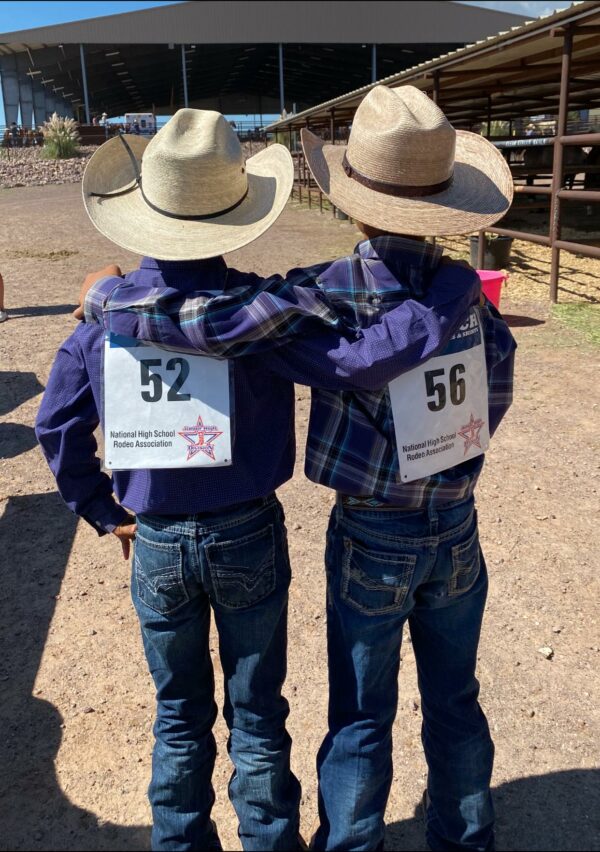 Twins wearing cowboy hats and outfits at a rodeo wearing numbers on their backs with arms around each other