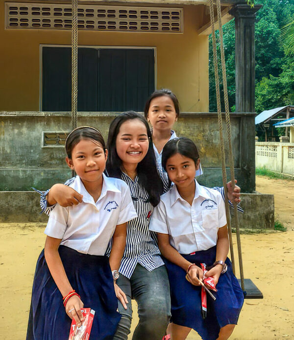 Young woman surrounded by 3 students in front of a Cambodian school