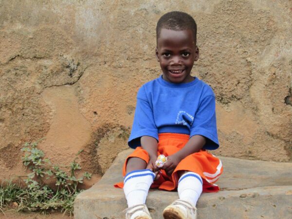 Little boy in a blue shirt and orange shorts sits on a wall, smiling