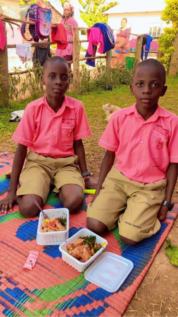 Twin boys in pink shirts and khaki shorts eating food on the ground in Uganda