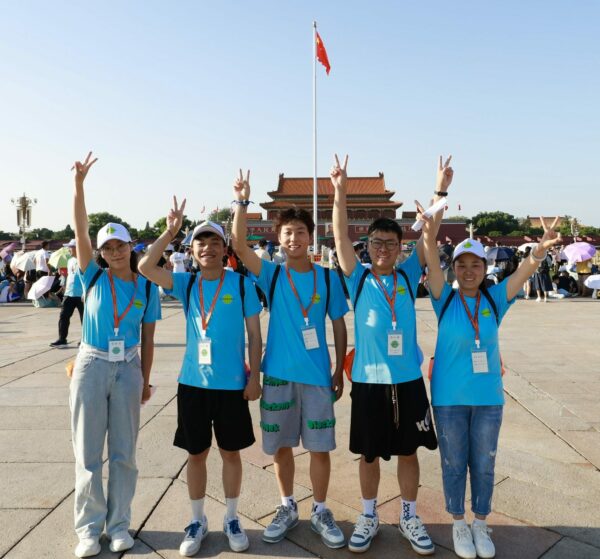5 young adults in matching blue shirts hold up their arms to give the peace sign