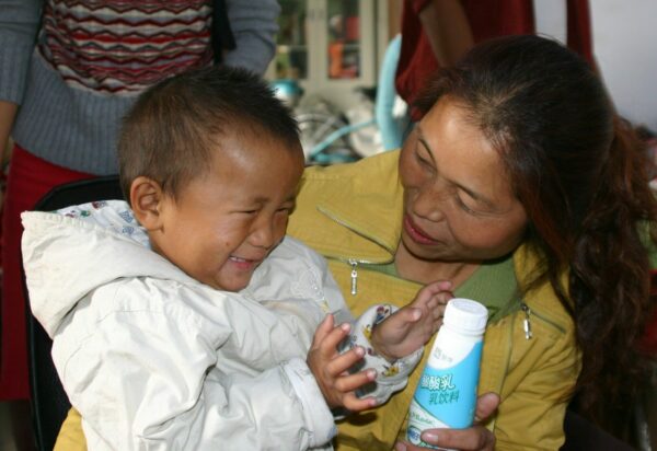 Little boy in a white jacket getting a drink from a woman wearing a gold jacket