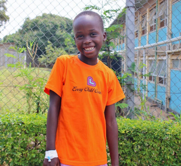 Ugandan girl in an orange shirt smiles in front of a hospital