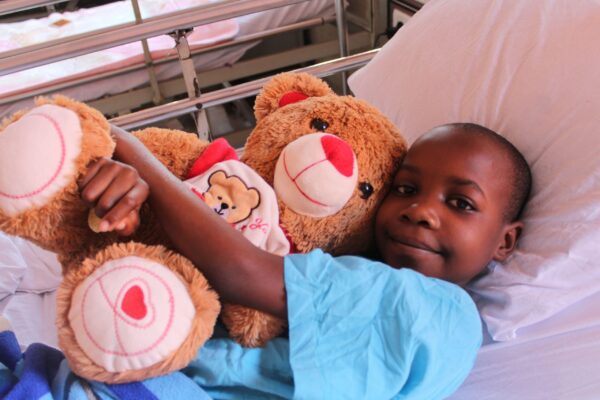 Girl pudding a big teddy bear while lying in hospital bed after cardiac surgery