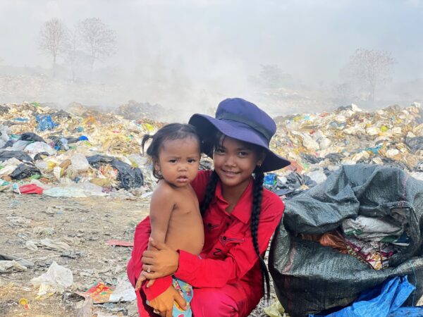 Girl in a blue hat holds her baby sister at a landfill in Cambodia