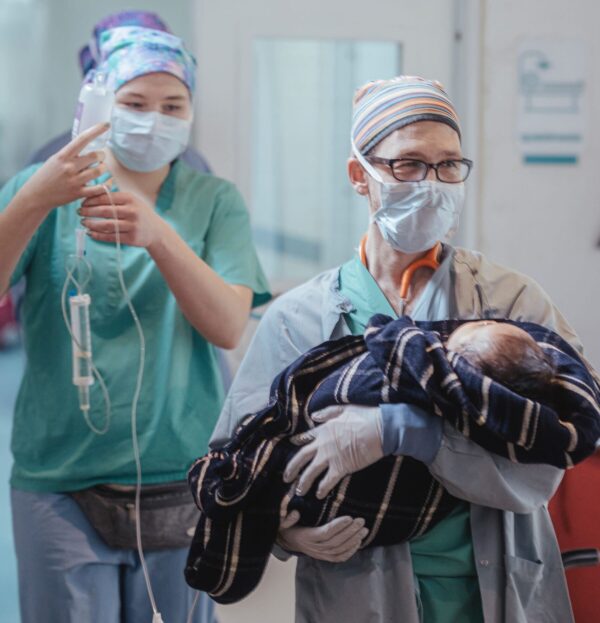 Doctor holds a baby in a blanket followed by a nurse holding an IV