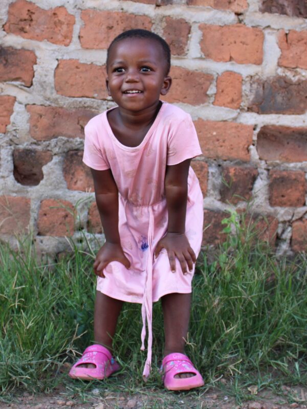 Little girl in a pink dress and shoes stands in front of a brick wall