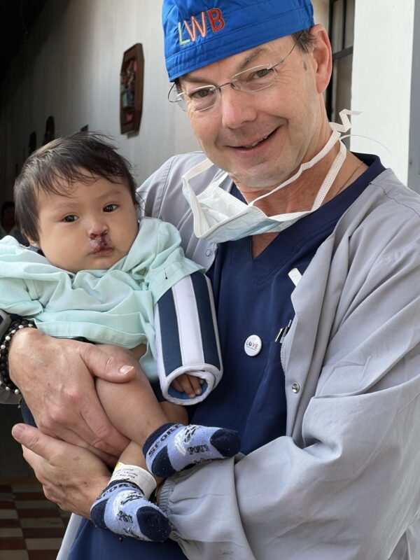 Man in surgical cap and scrubs holds a baby post cleft lip surgery